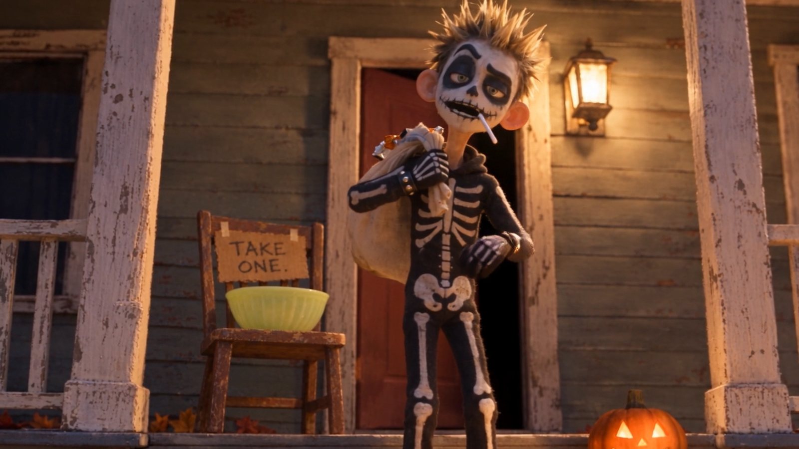A boy in a skeleton costume, a stuffed sack over his shoulder, on a porch beside a sign that says Take One and an empty bowl